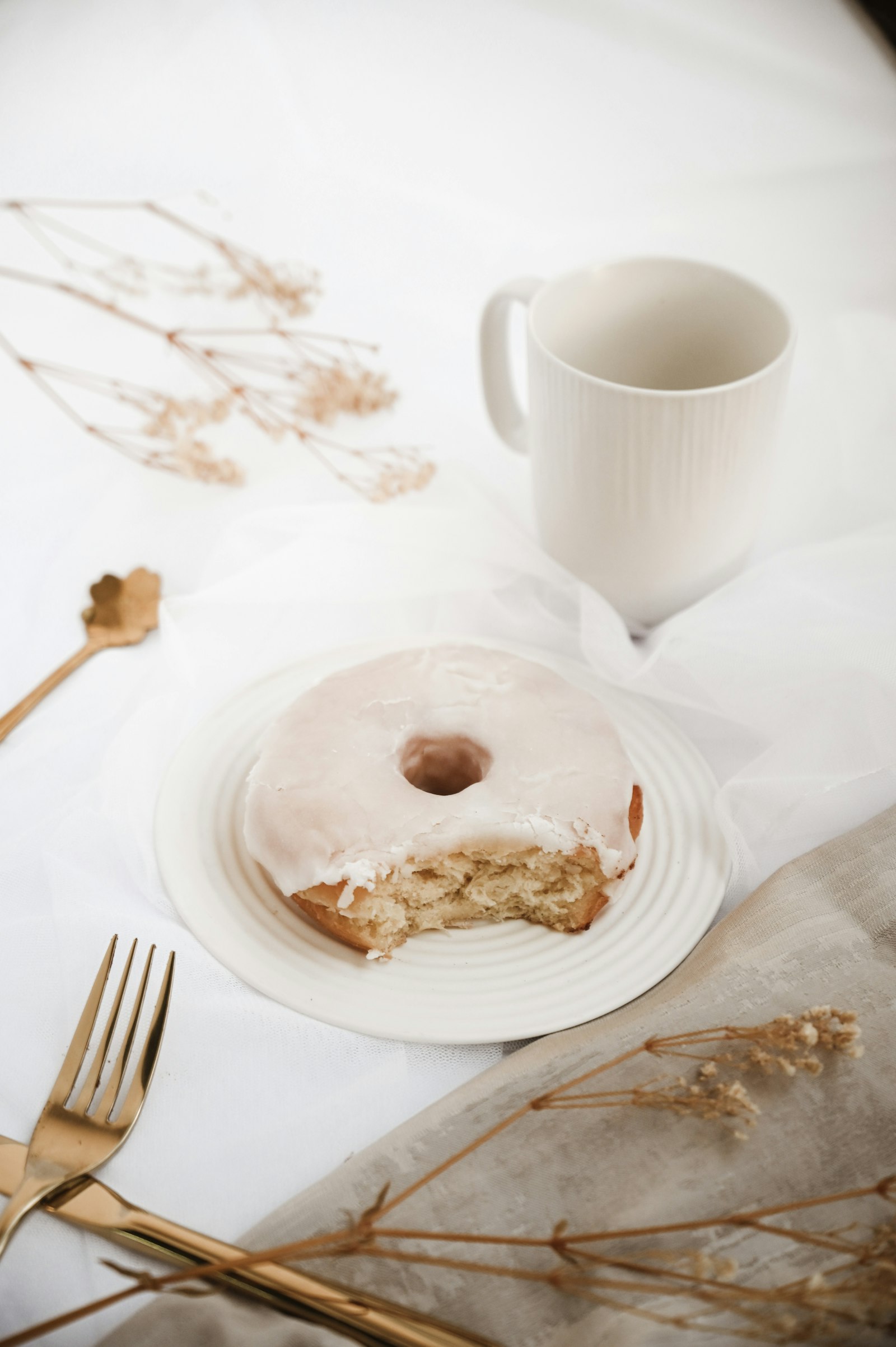 A glazed donut with a bite taken next to a white coffee mug in bright morning light. Representative stock photo.